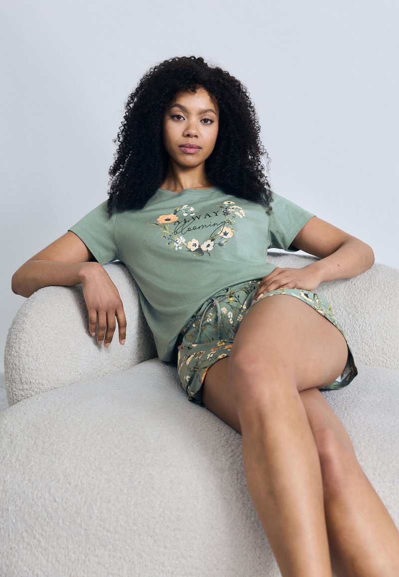 Πράσινο βαμβακερό t-shirt με floral σχέδιο και κείμενο, συνδυασμένο με μπλε σορτς με σχέδιο. Το μοντέλο κάθεται σε καναπέ με υφή και ανοιχτόχρωμο ύφασμα.