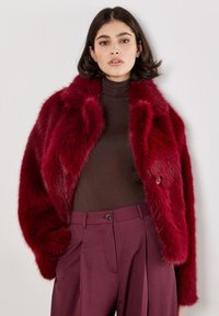 Burgunderfarbene Kunstfelljacke mit einer kastigen Form, doppelreihiger Gestaltung und plüschiger Textur. Kombiniert mit einem braunen Rollkragenpullover und lila Hosen.