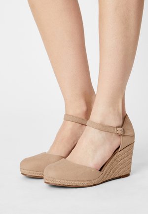 Beige suede gesloten neus plateau schoenen met enkelband en gesp, gedragen op blote voeten, met gevlochten touwdetail op de plateauhakken.