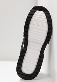 Semelle d'une basket avec un motif de bande de roulement noir et blanc appuyée contre un mur blanc et un sol blanc.