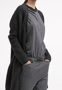 Lång svart cardigan över en grå zip-up topp och matchande joggers, med en avslappnad passform och dragsko i midjan, tillverkad av mjukt material.