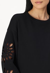 Schwarzer Pullover mit ausgeschnittenem Blumenmuster am linken Ärmel, mit Rundhalsausschnitt und weicher Textur. Leichter Stoff.