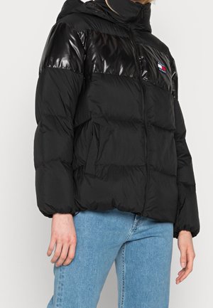 Czarna kurtka puchowa z błyszczącym górnym panelem i kapturem, z logo Tommy Jeans, noszona z jasnoniebieskimi dżinsami.
