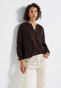 Vrouw met krullend haar die een donkerbruine V-hals blouse en gebroken witte broek draagt, staand tegen een witte muur met één hand in haar zak.