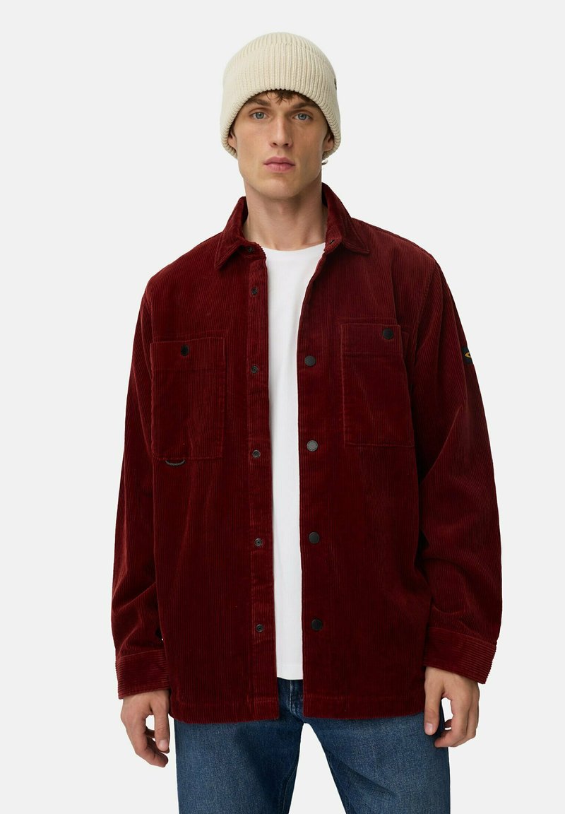 Ung mand iført en beige strikket hue, hvid t-shirt, rød corduroy skjortejakke og blå jeans, stående foran en ensfarvet baggrund.