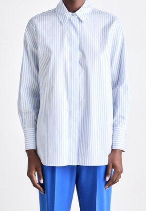 Personne portant une chemise à manches longues rayée bleu clair et blanche avec des boutons et un pantalon bleu, debout devant un fond uni.