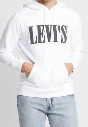 Osoba ubrana w białą bluzę z kapturem Levi's z dużym czarnym logo i jasnoniebieskie jeansy, ręce w przedniej kieszeni bluzy.