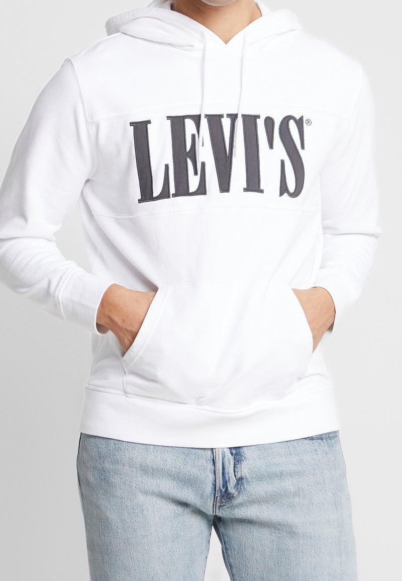 Personne portant un sweat à capuche blanc Levi's avec un grand logo noir et un jean bleu clair, les mains dans la poche avant du sweat.