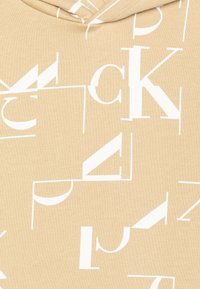 Beige Kapuzenpullover aus strukturiertem Stoff mit großen, unregelmäßig platzierten weißen Buchstaben und Akzenten, die ein modernes, grafisches Muster erzeugen.