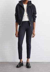 Svart zip-up-hoodie med luva, bärs över en vit topp; kombinerad med åtsittande svarta byxor och kraftiga svarta loafers. Slätt tyg och skräddarsydd passform.