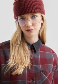 Jonge vrouw met blond haar, het dragen van gouden brillen, een rode gebreide muts en een rood-grijze geruite blouse met een zwarte kraag.