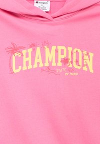 Ροζ ανοιχτόχρωμο φούτερ με κουκούλα, με το "CHAMPION" έντονα κίτρινο και διακοσμητικά γραφικά με φοίνικες και ήλιο, κατασκευασμένο από απαλό βαμβακερό ύφασμα.