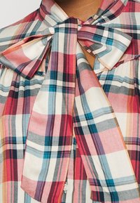 Geruite blouse met een strik bij de halslijn. Kleuren zijn rood, roze en blauw op een lichte achtergrond. Gladde stof met een gestructureerd ontwerp.