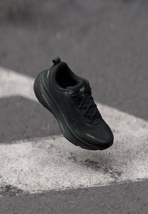Zapatilla de correr negra Nike en el aire sobre el asfalto con una marca vial blanca, destacando su diseño ligero y el movimiento.