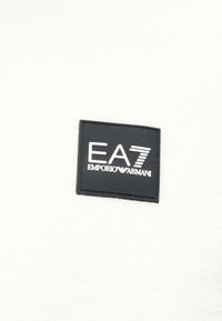 Eticheta rectangulară neagră din piele cu textul embosat "EA7 EMPORIO ARMANI" pe un fundal moale din material alb.