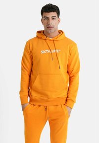 Sudadera naranja con capucha, cordones ajustables, bolsillo frontal y logo blanco "SIXTHJUNE". Combinada con pantalones de chándal a juego.