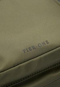 Mračna zelena platnena ruksak s glatkom teksturom i istaknutim plavim logotipom "PIER ONE". Detaljno šivenje duž šavova.