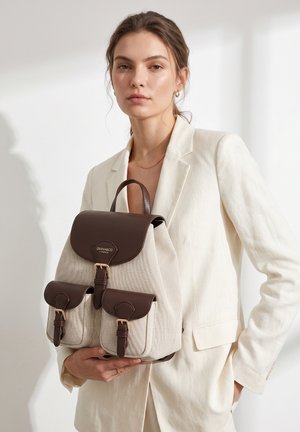 Woman in cream blazer holding beige canvas backpack with dark brown leather flaps and buckles, standing against light background.