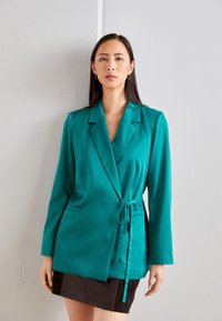 Blazer turquoise à manches longues, avec une ceinture �à nouer et des revers crantés ; texture lisse, doté de deux poches latérales et d'une coupe structurée.