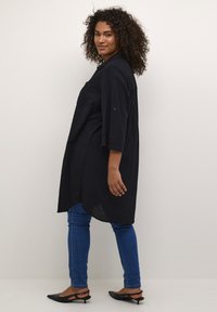 Tunique noire, longueur genoux, en tissu texturé, à manches trois-quarts, associée à un denim bleu foncé et des ballerines noires à bout pointu.