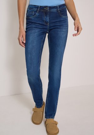 Kvinde iført mørkeblå slim-fit jeans og tan slip-on sko med spænder, stående mod en ensfarvet lys baggrund.