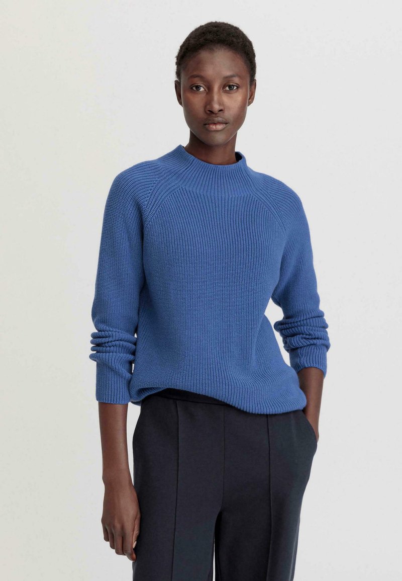 Pull en tricot bleu avec un col montant, texture côtelée et manches longues. Associé à un pantalon tapered sombre.