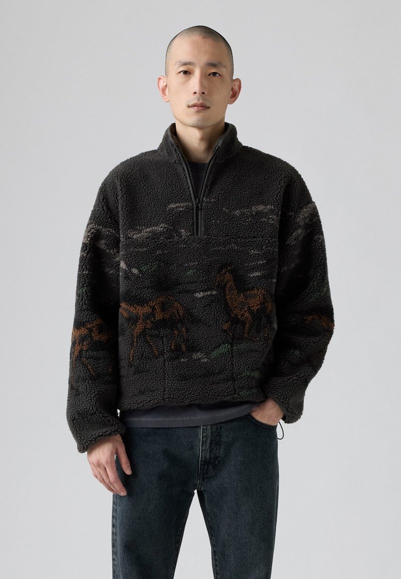 Mann in einem schwarzen Fleece-Pullover mit braunen Giraffenmustern, der mit einer Hand in der Tasche vor einem schlichten grauen Hintergrund steht.