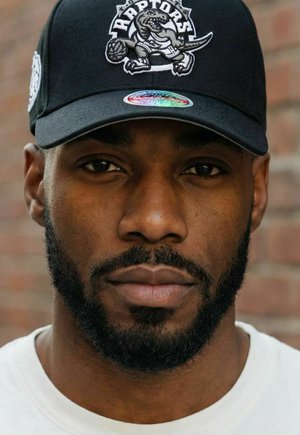 Mann mit gestutztem Bart, der eine schwarze Raptors-Baseballkappe und ein weißes Hemd trägt, nach vorne blickend vor einem verschwommenen Hintergrund aus einer Backsteinwand.