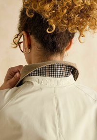 Jachetă de culoare crem cu guler din catifea, având un căptușel cu carouri. "JOULES" este gravat pe spate. Părul ondulat este vizibil.