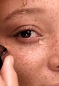 Gros plan du visage d'une personne appliquant du fond de teint avec une éponge de maquillage, présentant une peau couleur brun clair, des taches de rousseur et des yeux marron.