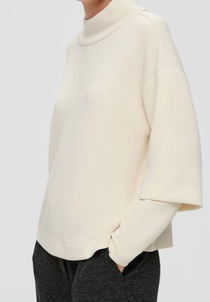 Person trägt einen cremefarbenen, geschichteten Pullover mit weiten, kurzen Ärmeln über langen Ärmeln, kombiniert mit dunkler, strukturierter Hose.