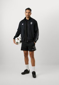 Jachetă sport neagr�ă cu logo și dungi albe, purtată cu pantaloni scurți negri. Ținând o minge de fotbal cu accente verzi și negre, purtând ghete negre.