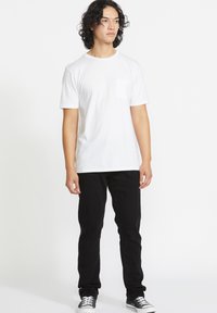 Tricou alb din bumbac cu mâneci scurte și un buzunar pe piept, asortat cu blugi negri și adidași negri, stând în fața unui fundal alb.