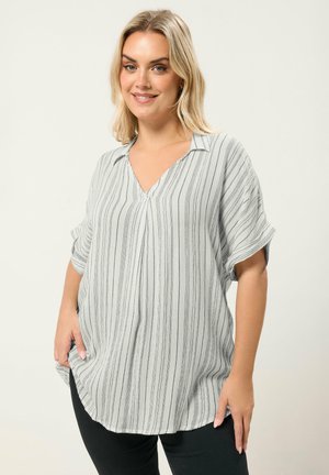 Lyshåret kvinde iført en løs, hvid V-hals top med lodrette sorte striber og sorte bukser, smilende med hænderne i lommerne.