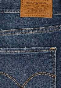 Blå denimjeans har en läderetikett med varumärkestext och detaljer. Kontrastsydd sticker ut vid fickorna och sömmarna, vilket tillför struktur.