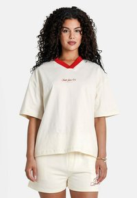 Camiseta de algodón blanca con escote en V rojo y logotipo bordado, combinada con pantalones cortos de algodón blancos que cuentan con un pequeño detalle bordado.