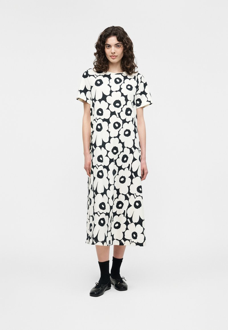 Γυναίκα με σγουρά μαλλιά που φοράει μαύρο και άσπρο midi φόρεμα με μεγάλο floral μοτίβο, μαύρες κάλτσες και μαύρα δερμάτινα παπούτσια με κορδόνια, στέκεται μπροστά σε λευκό φόντο.