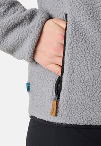 Grå fleecejacka med texturerat material, ficka med blixtlås och läderdragband. Inkluderar en flaggdetalj på ärmen.