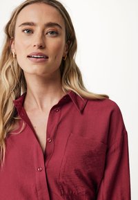 Luftige, bordeauxfarbene Bluse mit Kragen, Fronttasche und Knopfverschluss. Weicher, strukturierter Stoff mit sichtbaren Nähdetails.