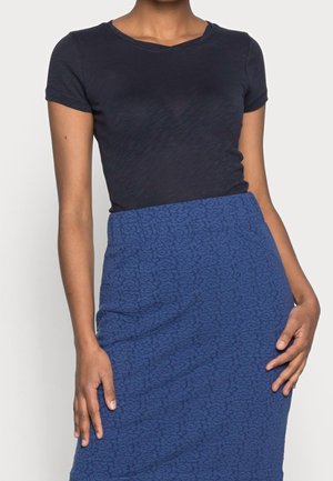 Femme portant un t-shirt ajusté à manches courtes bleu marine foncé rentré dans une jupe bleue texturée taille haute, debout les mains près des hanches.