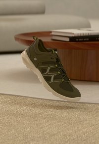 Σκούρο πράσινο αθλητικό παπούτσι με ανοιχτόχρωμη σόλα και κίτρινες λεπτομέρειες, τοποθετημένο πάνω από μπεζ χαλί, ξύλινο στρογγυλό τραπέζι και στοίβα βιβλίων σε σαλόνι.