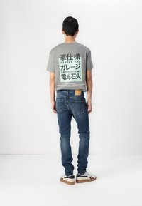 Camiseta gris con un gran texto verde en la parte posterior, combinada con jeans azules y zapatos blancos con acentos negros.
