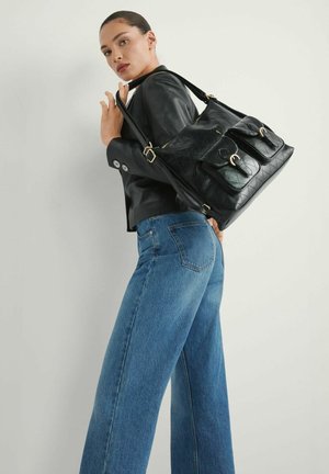 Kobieta w czarnej skórzanej kurtce i niebieskich dżinsach, niosąca na ramieniu dużą czarną pikowaną torebkę z dwoma przednimi kieszeniami.