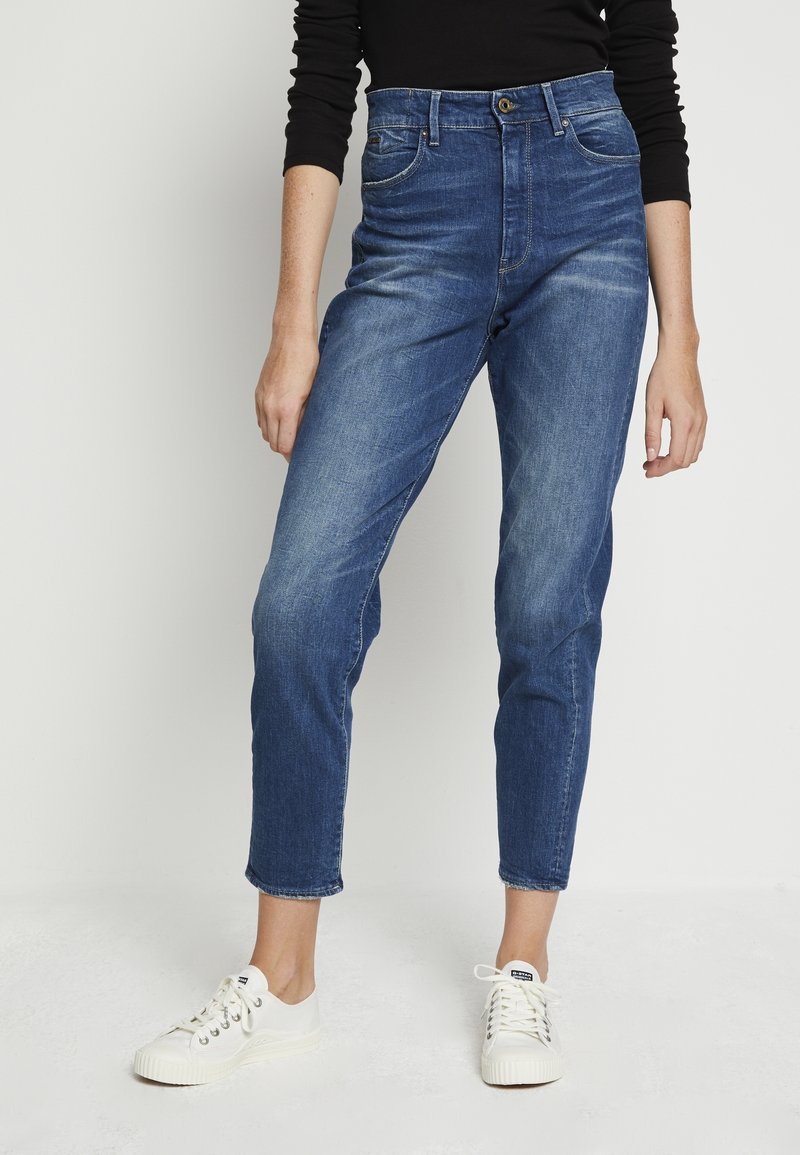 Jean bleu taille haute avec décoloration légère, cinq poches et fermeture à bouton, associé à des baskets blanches sur un fond uni.