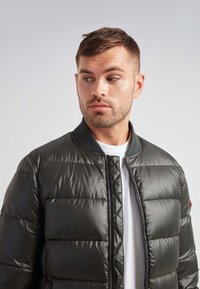 Giubbotto imbottito di colore verde scuro, con zip frontale, colletto a coste e pannelli cuciti. Indossato sopra una camicia bianca.