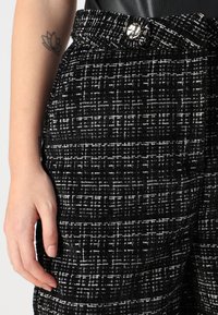 Pantalon en tweed noir et blanc avec un motif texturé, doté d'une fermeture à bouton et de poches latérales visibles. Conçu pour une coupe ajustée.
