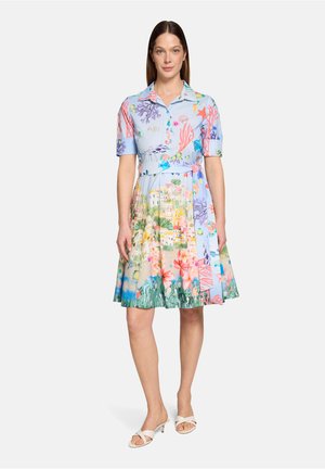 Kvinde står iført en kjole med knapper og korte ærmer med farverige mønstre af havliv og kystby, kombineret med hvide sandaler med hæl.