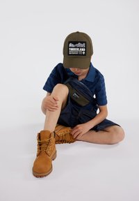 Bottes en cuir marron avec des lacets jaunes, shorts en denim et polo bleu à motif. Une casquette verte arbore un graphique de montagne et un logo.