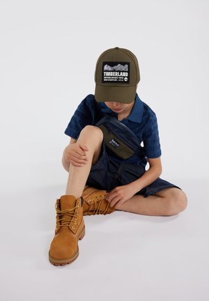 Bottes en cuir marron avec des lacets jaunes, shorts en denim et polo bleu à motif. Une casquette verte arbore un graphique de montagne et un logo.