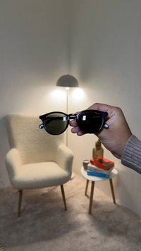 Lunettes de soleil noires à monture œil-de-chat avec des verres foncés et des accents métalliques, tenues par une main sur un fond neutre comprenant une chaise et une lampe.